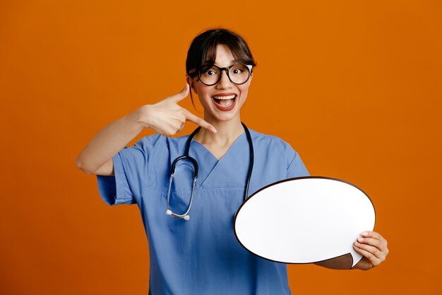 Pleased holding and points at speech bubble young female doctor wearing uniform fith stethoscope isolated on orange background