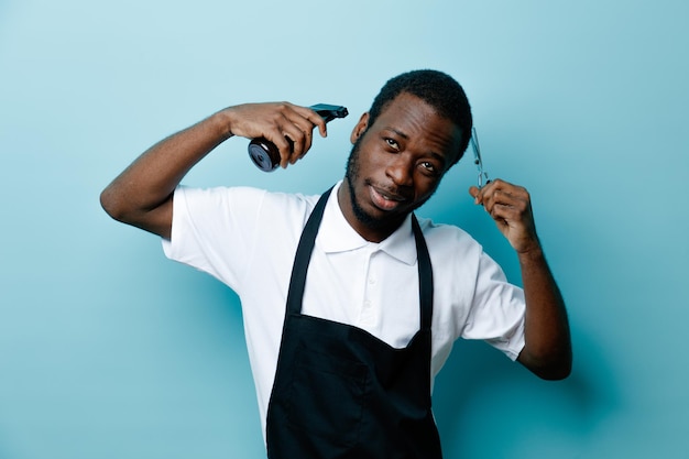 Pleased holding spray bottle with scissors young african american barber in uniform isolated on blue background