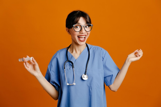 Felice azienda siringa giovane dottoressa che indossa uno stetoscopio fith uniforme isolato su sfondo arancione