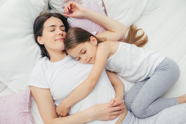 Pleased little child sleeps near her mother embraces with love\
has pleasant dreams lie on comfortable bed mum and cute daughter\
have good sleep in bedroom family sleeping and rest concept