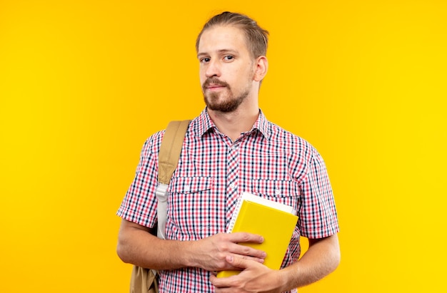 Pleased looking camera young guy student wearing backpack holding book isolated on orange wall