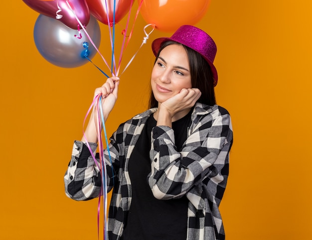 Pleased looking side young beautiful girl wearing party hat holding balloons putting hand on cheek 