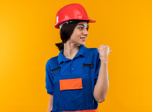 Pleased looking at side young builder woman in uniform points at behind isolated on yellow wall with copy space