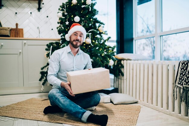 довольный мужчина сидит возле елки и разворачивает картонную коробку с подарком на Рождество
