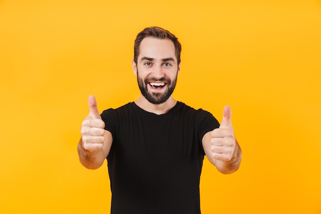 pleased man wearing basic black t-shirt smiling and gesturing thumbs up isolated over yellow wall