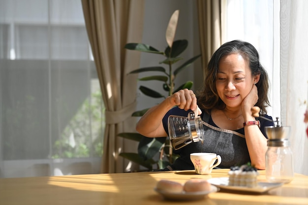 Pleased middle aged sitting in bright living room brewing drip coffee and enjoys her morning