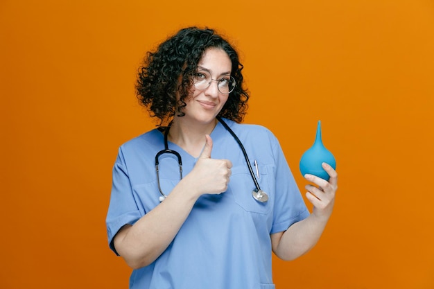 pleased middleaged female doctor wearing uniform glasses and stethoscope around her neck showing enema looking at camera showing thumb up isolated on orange background