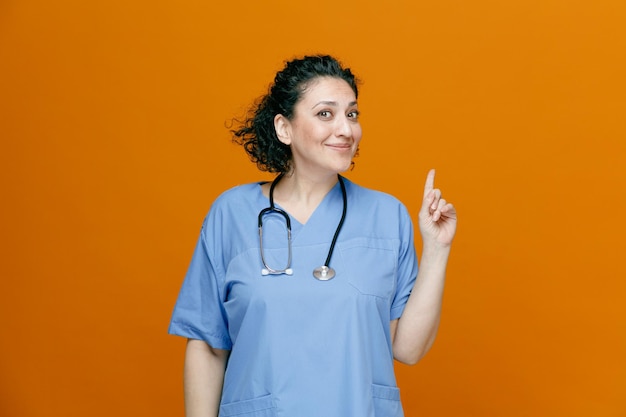 Pleased middleaged female doctor wearing uniform and stethoscope around her neck looking at camera pointing up isolated on orange background