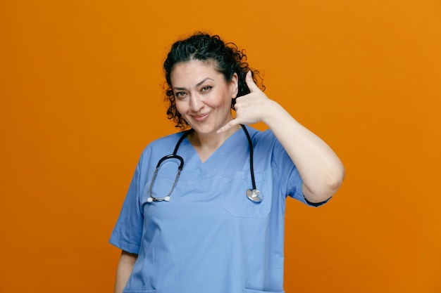 Довольная женщина-врач средних лет в униформе и со стетоскопом на шее смотрит в камеру, показывающую жест вызова, изолированный на оранжевом фоне