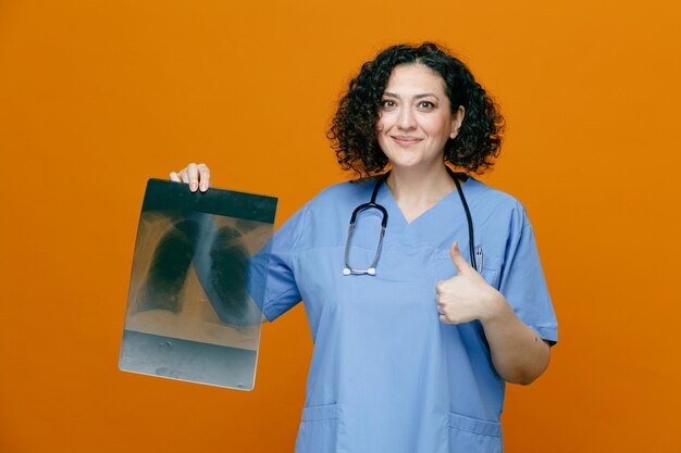 Pleased middleaged female doctor wearing uniform and stethoscope around her neck looking at camera showing xray shot and thumb up isolated on orange background