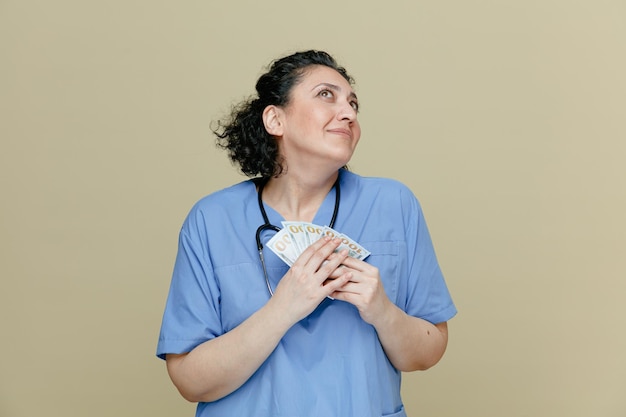 pleased middleaged female doctor wearing uniform and stethoscope around neck holding money with both hands looking up isolated on olive background