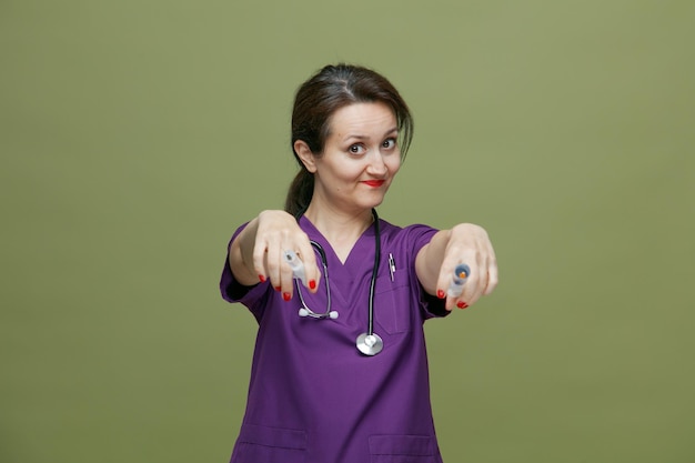 Felice dottoressa di mezza età che indossa uniforme e stetoscopio intorno al collo guardando la telecamera allungando siringhe con aghi verso la telecamera isolata su sfondo verde oliva