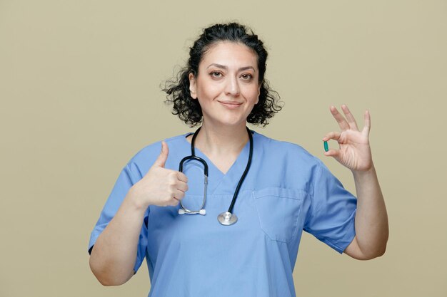 pleased middleaged female doctor wearing uniform and stethoscope around neck showing capsule looking at camera showing thumb up isolated on olive background