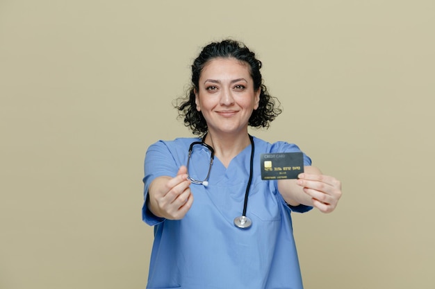 Pleased middleaged female doctor wearing uniform and stethoscope around neck stretching credit card out towards camera looking at camera making money gesture isolated on olive background