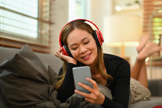 Photo pleased millennial woman with wireless headphone lying on comfortable couch and using smart phone