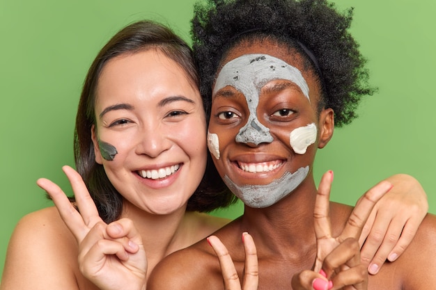Pleased multiethnic young women stand closely to each other have fun undergo beauty procedures apply clay masks