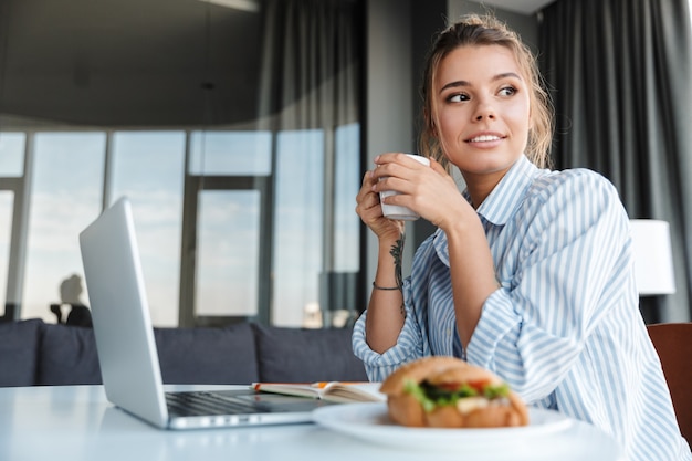 リビングルームのテーブルに座ってコーヒーを飲み、ラップトップを使用して喜んでいる素敵な女性