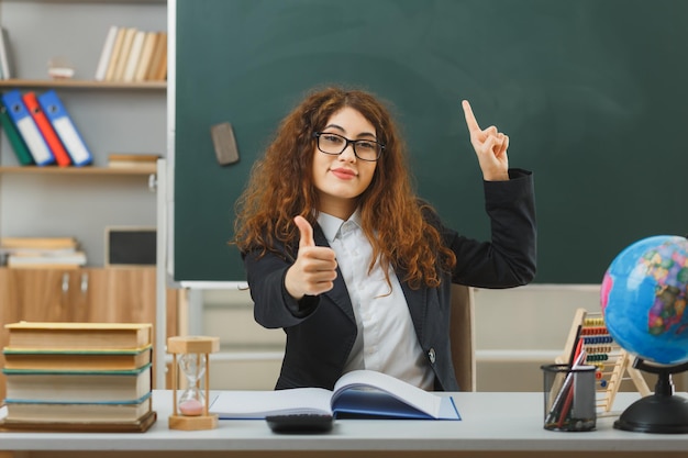 pleased points aat up showing thumbs up young female teacher wearing glasses sitting at desk with school tools in classroom