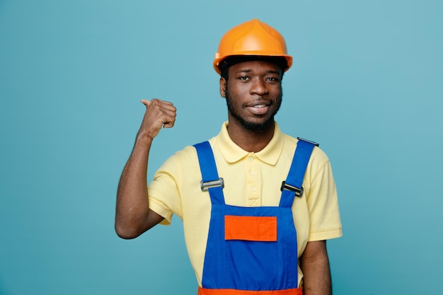 Pleased points at back young african american builder in uniform isolated on blue background