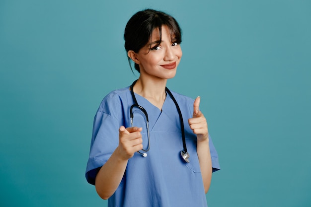 Piacere punti alla fotocamera giovane dottoressa che indossa uno stetoscopio fitth uniforme isolato su sfondo blu