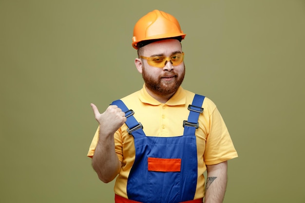 Pleased points at side young builder man in uniform isolated on green background