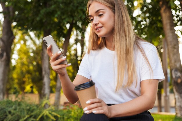 Pleased positive optimistic young lady sit in nature park using mobile phone drinking coffee.