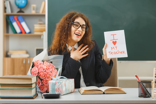 Photo pleased putting hand on body young female teacher holding postcard sitting at desk with school tools in classroom