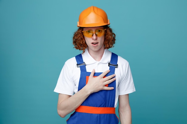 pleased putting hand on heart young builder man in uniform isolated on blue background