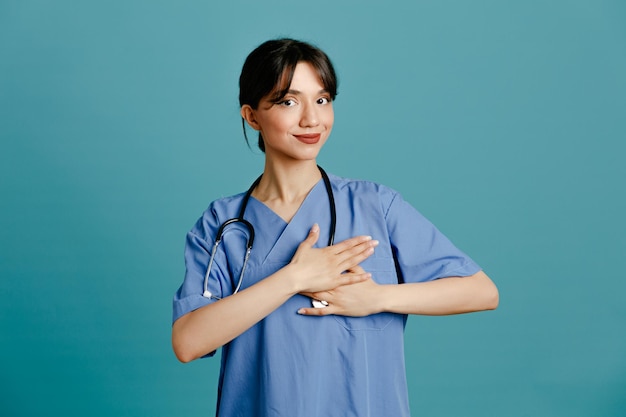 Piacere di mettere la mano sul cuore giovane dottoressa che indossa uno stetoscopio fitth uniforme isolato su sfondo blu