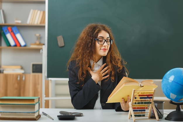 Felice di mettere la mano sul cuore giovane insegnante femminile che indossa occhiali tenendo e leggendo un libro seduto alla scrivania con strumenti scolastici in classe