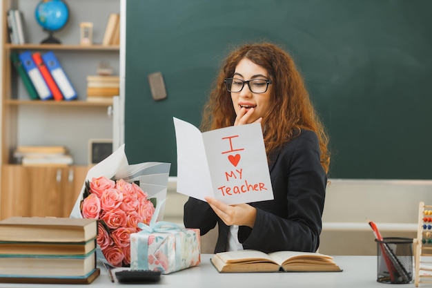 pleased putting hand on mouth young female teacher holding postcard sitting at desk with school tools in classroom