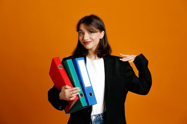 Pleased putting hand on shoulder holding folders young beautiful female wearing black jacket isolated on orange background