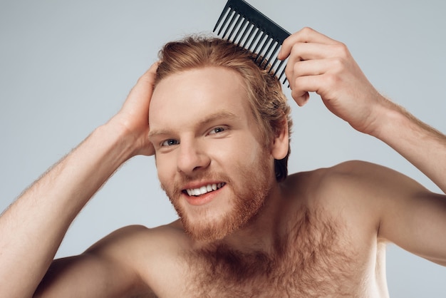 Pleased red haired man combs hair with comb.