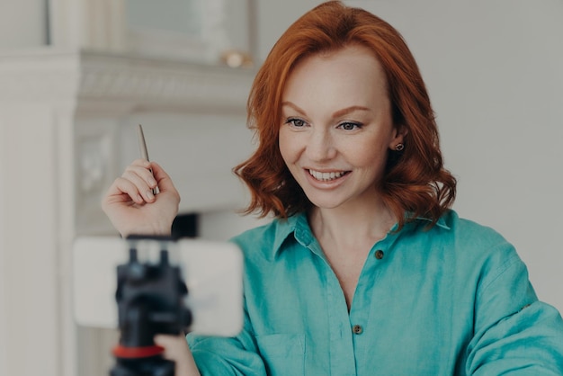 Pleased redhead young European woman uses front cellphone camera on tripod has personal vlog shoots video gives some recommendations dressed in shirt talks with followers Blogging concept