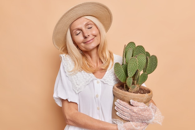 Pleased relaxed woman with fair hair keeps eyes closed holds pot of green prickly succulent cactus wears hat white dress lacy gloves feels satisfied gets houseplant as present isolated over beige wall