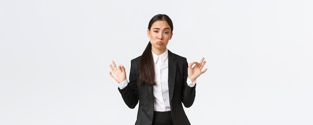 Pleased sassy asian businesswoman in black suit show not bad gesture nod in approval and make okay gesture very good work praising coworker standing satisfied over white background