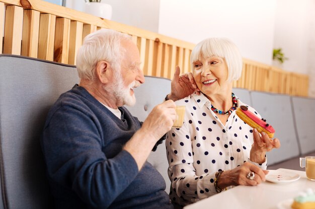 Pleased senior woman holding eclair and senior man drinking coffee