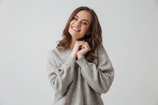 Pleased sensual brunette woman in sweater holding arms together  over grey wall