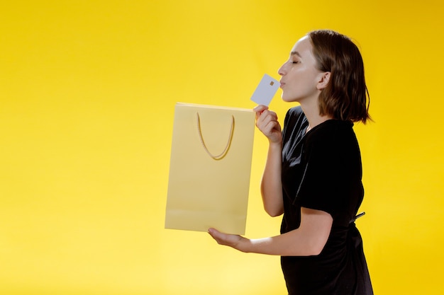 Pleased shopaholic woman posing shopping bags and credit card on yellow background.