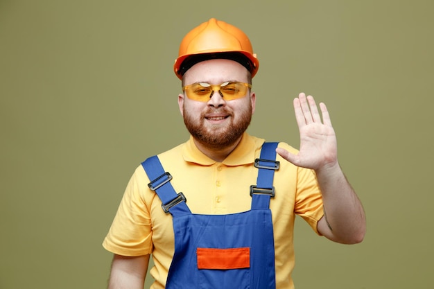 Pleased showing hello gesture young builder man in uniform isolated on green background
