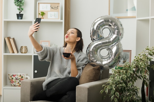 Pleased showing kiss gesture beautiful woman on happy women day holding glass of wine take a selfie sitting on armchair in living room