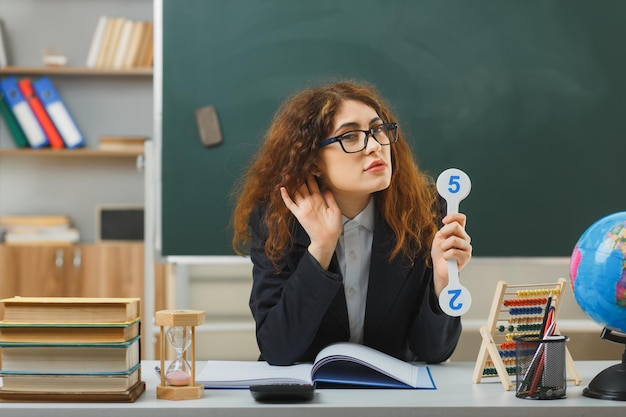 教室で学校の道具を持って机に座っている数のファンを保持している眼鏡をかけている若い女性教師が聞くジェスチャーを見せて喜んで