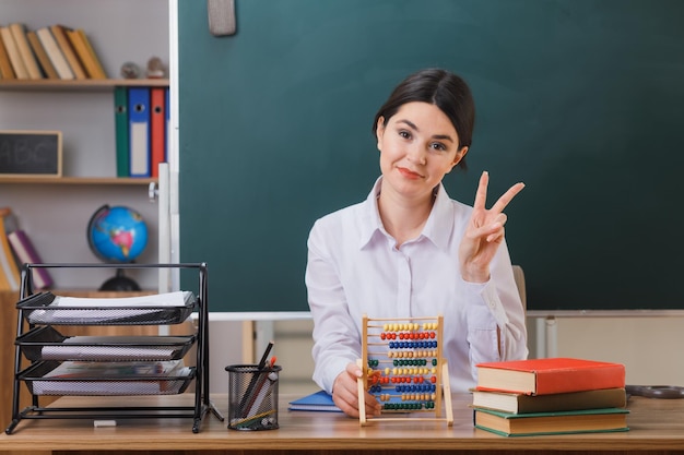 pleased showing peace gesture young female teacher holding abacus sitting at desk with school tools in classroom