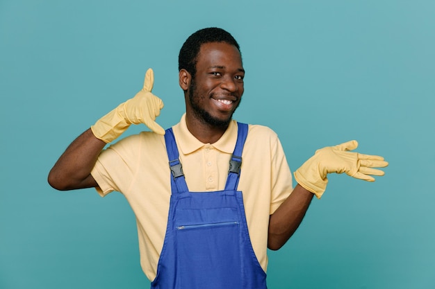 青の背景に分離された手袋と制服を着た若いアフリカ系アメリカ人クリーナー男性を示す電話ジェスチャーを喜んで