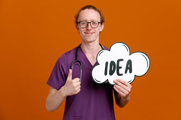 Pleased showing thumbs up holding idea bubble young male doctor wearing uniform with stethoscope isolated on orange background