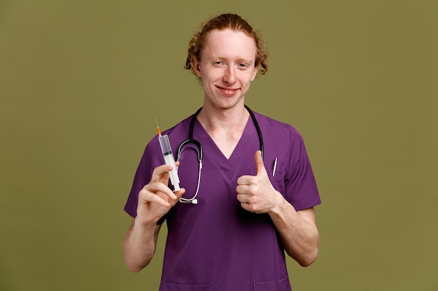 Pleased showing thumbs up young male doctor wearing uniform with stethoscope holding syringe isolated on green background