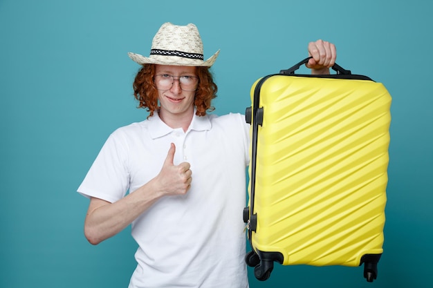 Приятно показывать большой палец вверх молодому путешественнику в шляпе и очках, держащим чемодан на синем фоне