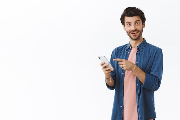 Pleased smiling handsome man with beard in pink t-shirt, shirt, holding smartphone, pointing screen and grin camera as recommend friend log in and try yourself