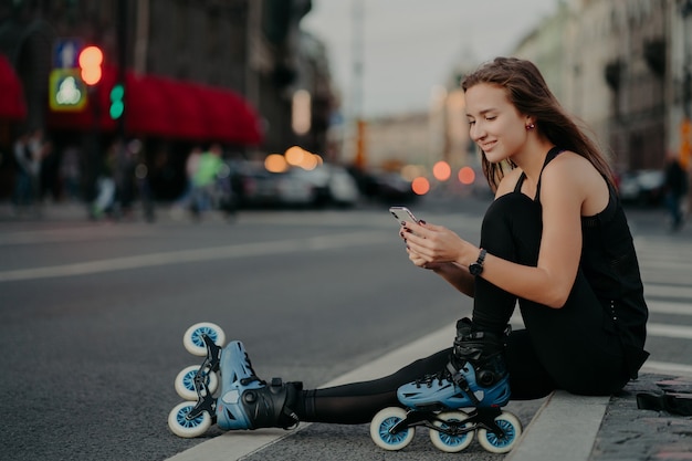 Pleased sporty woman wears sportsclothes rollerblades sits on road checks newsfeed via smartphone takes break after inline skating poses against blurred city background engaged in healthy lifestyle