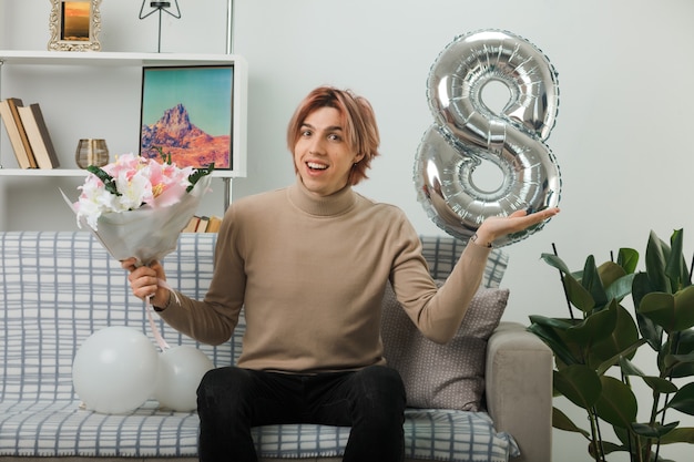 Pleased spreading hand handsome guy on happy women day holding bouquet sitting on sofa in living room